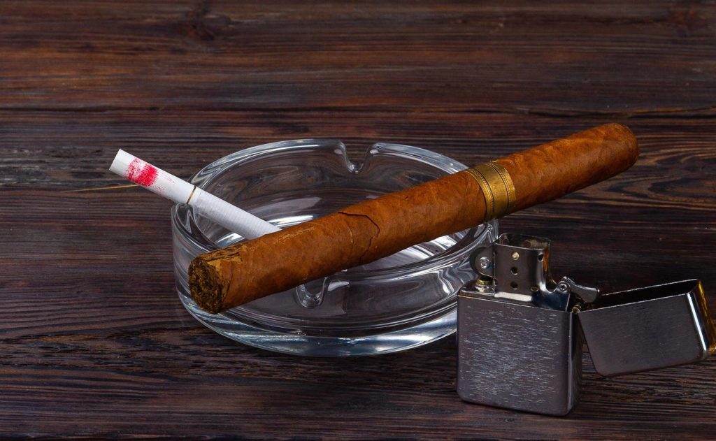 A cigar and cigarette with lipstick in an ashtray, next to a lighter, promoting a cigarette shop Tulsa.