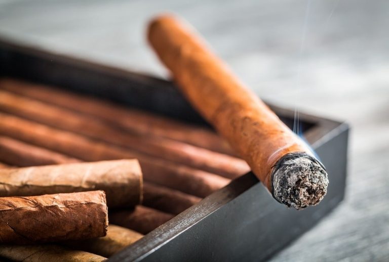 Close-up of lit cigar with ash and smoke on wooden tray, highlighting luxury tobacco from Cigar Shop Tulsa premium collection.