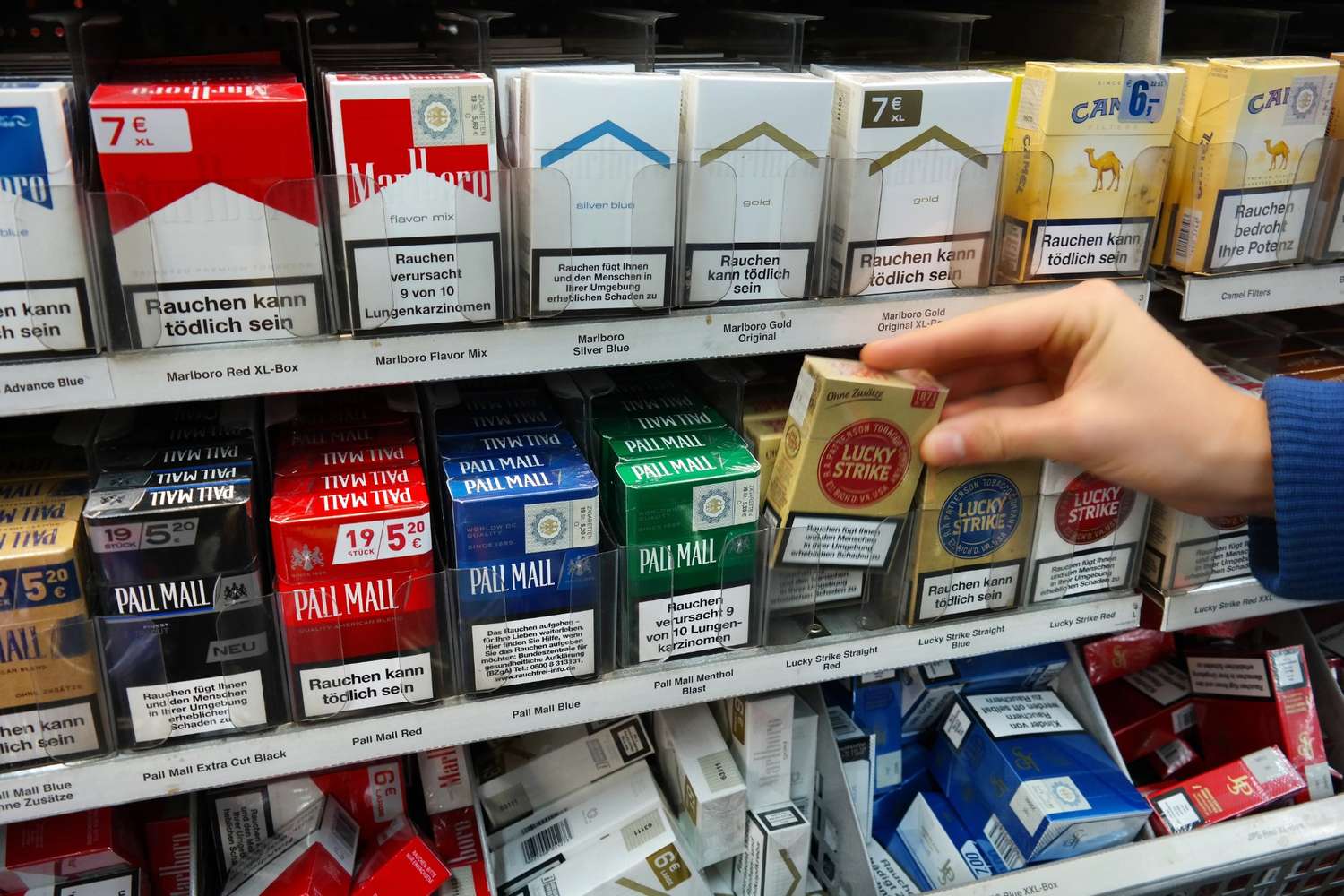 Hand selecting a Lucky Strike pack from a cigarette shop Tulsa shelf, displaying diverse brands like Marlboro and Pall Mall.