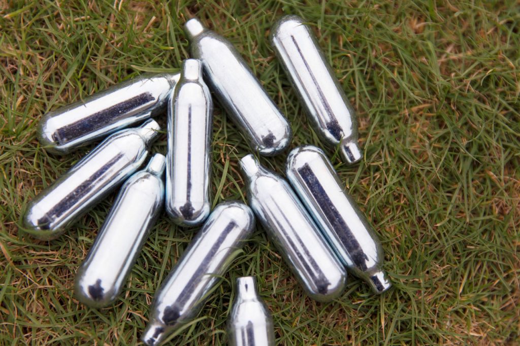 A pile of empty silver cream canisters showing used nitrous oxide chargers Tulsa litter found outdoors on green grass.