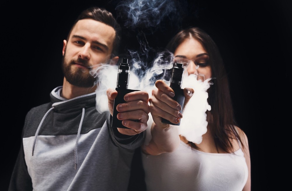 Man and woman hold out popular vape mods with thick clouds, showing top-rated e-cigarettes Tulsa options.