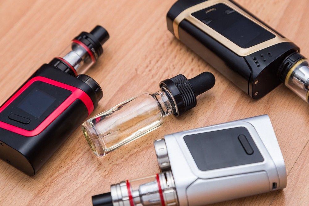 A black electronic cigarette device rests on a wooden table beside a full glass bottle from a vape store Tulsa stock.