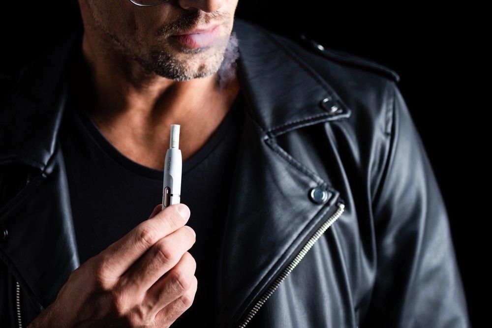 Bearded man in a leather jacket holding a slender white e-cigarette with visible vapor, exploring e-cigarettes tulsa options.