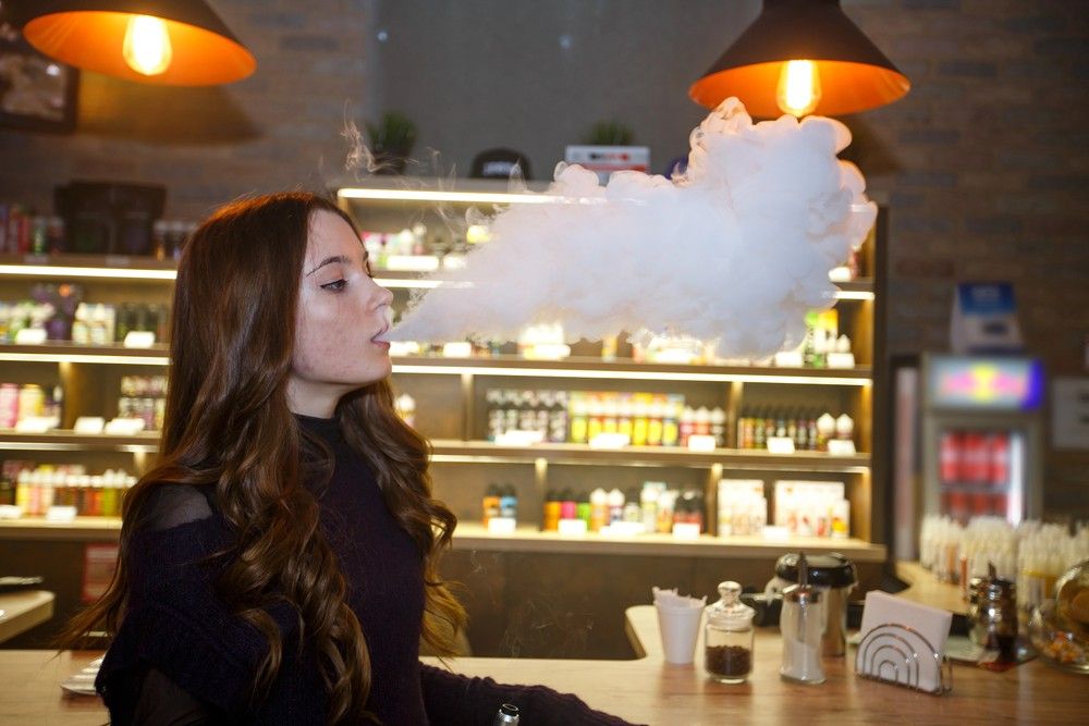 Long haired woman blowing a large vapor cloud inside a modern vape store Tulsa shop with illuminated juice displays.